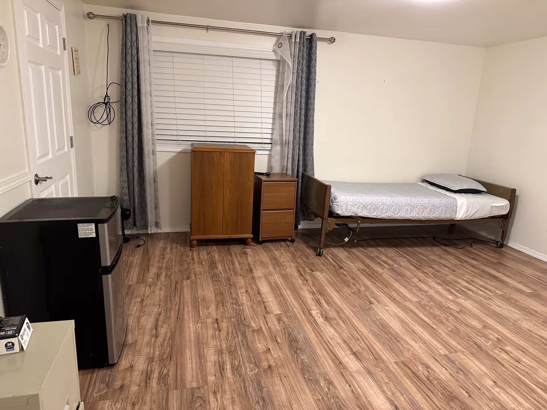 A simple bedroom with a single bed on wheels, a pillow and blanket neatly arranged. There is a wooden cabinet and a small wooden drawer unit next to a window with closed white blinds and gray curtains. A small black refrigerator is placed near a closed white door. The floor is covered with wood-patterned laminate.