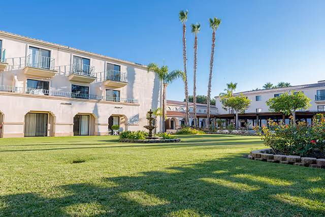 A sunny outdoor courtyard area at Brookdale San Marcos featuring a well-maintained green lawn, a central fountain, tall palm trees, and surrounding two-story buildings with balconies and windows.