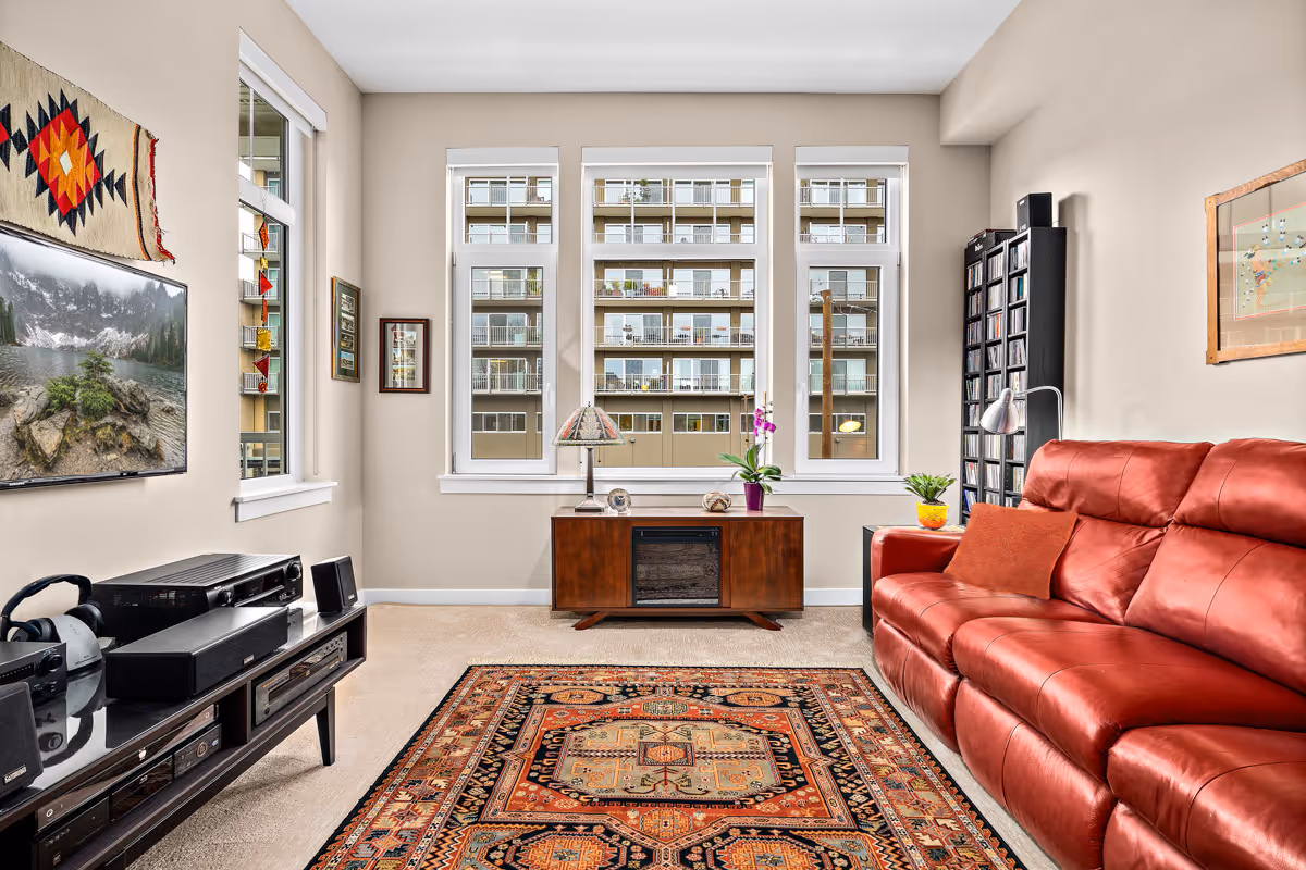 Bright living room with a red leather sofa, patterned rug, TV console, and three large windows overlooking an adjacent building.
