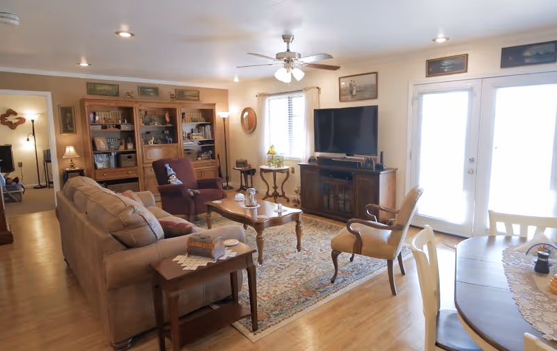A cozy living room with a beige sofa, a maroon armchair, a wooden coffee table on a patterned rug, and a TV on a wooden stand. There is a ceiling fan with lights, framed pictures on the walls, a window with curtains, and glass doors letting in natural light. A round dining table with chairs is partially visible on the right side.