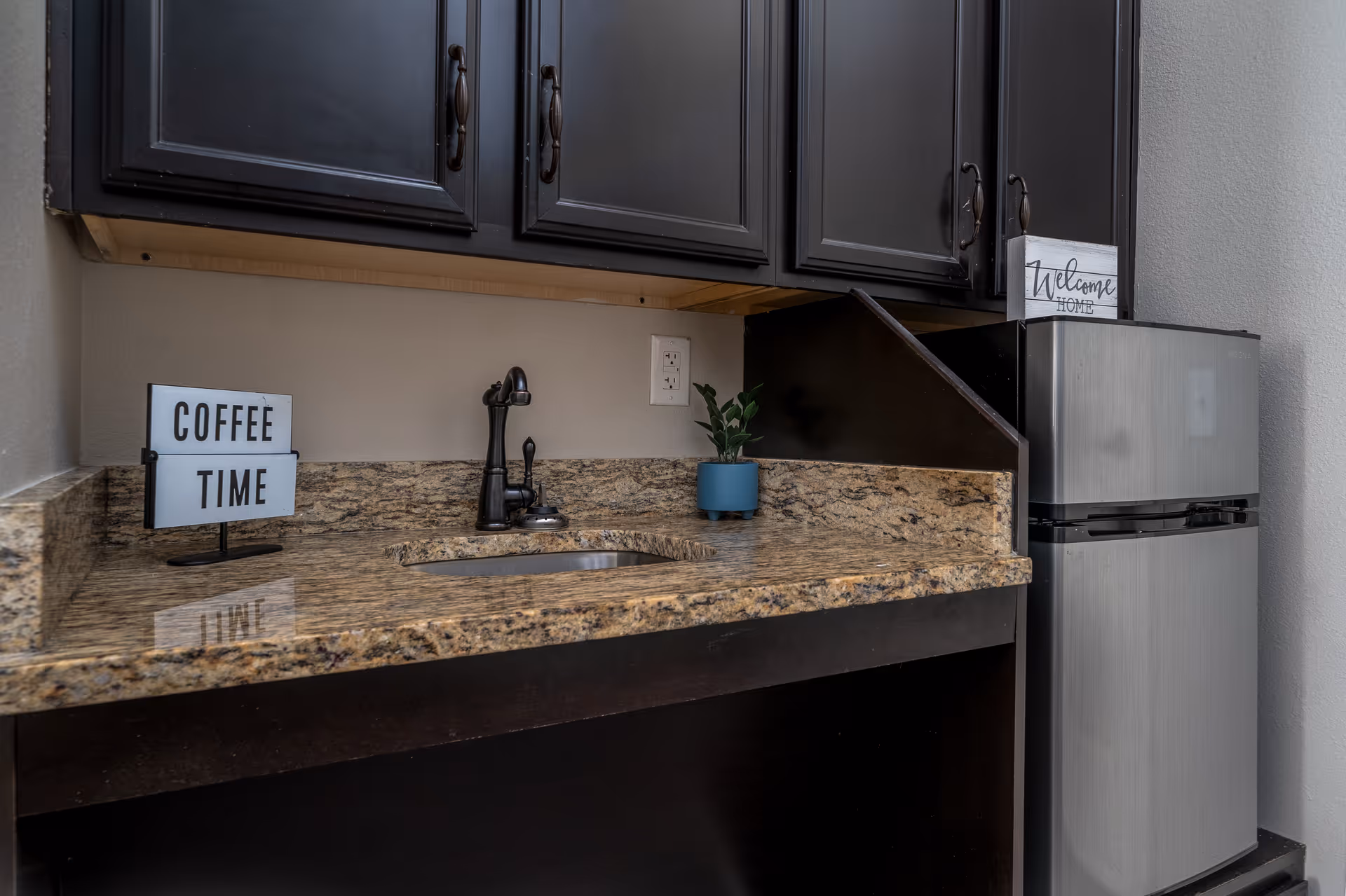 A small kitchen area with dark cabinets, a granite countertop with an inset sink and black faucet, a small potted plant, a mini refrigerator, and decorative signs that say 'COFFEE TIME' and 'Welcome HOME'.