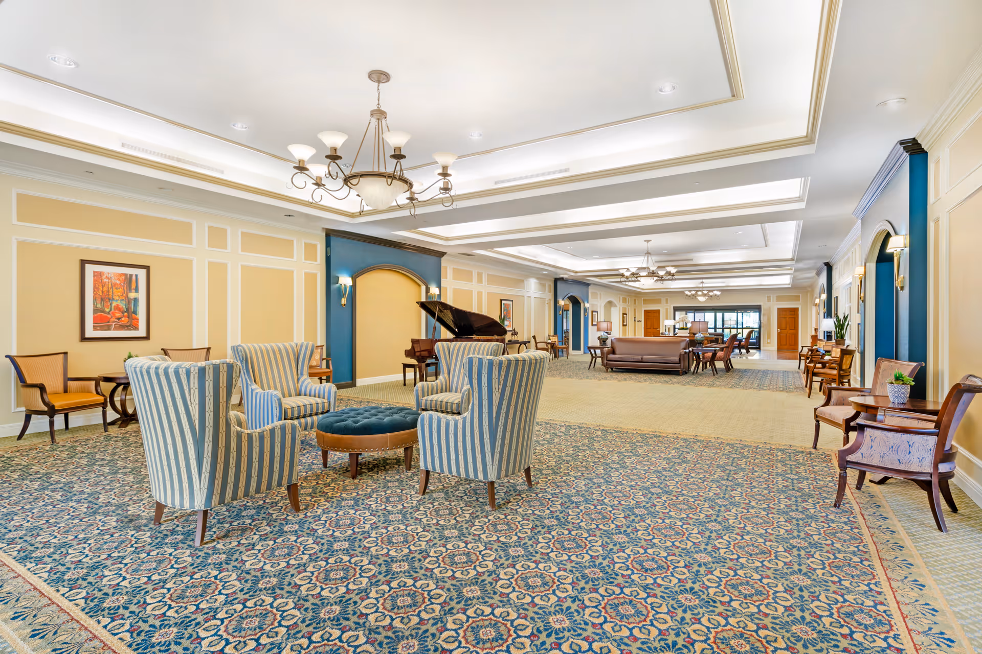 Spacious furnished common lounge with patterned carpet, grouped upholstered chairs, a grand piano, chandeliers, and additional seating areas.