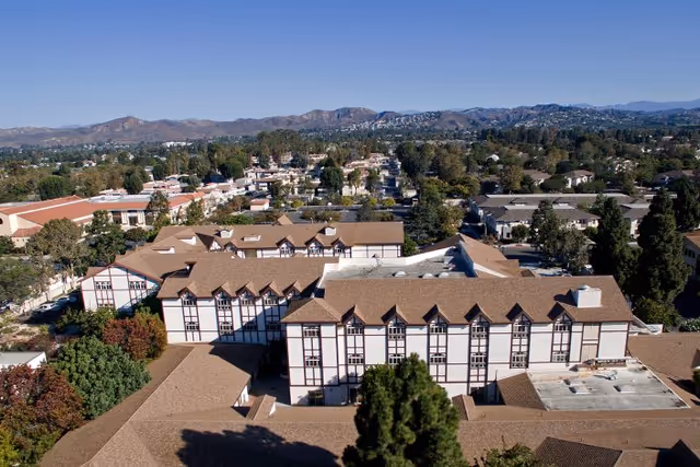 Aerial view of Lexington Assisted Living facility showing multiple connected buildings with brown roofs and white walls with dark trim, surrounded by trees and residential neighborhood with mountains in the background under a clear blue sky.