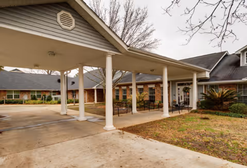 Covered entrance with white columns leading to a brick building facade, outdoor seating, and landscaping.