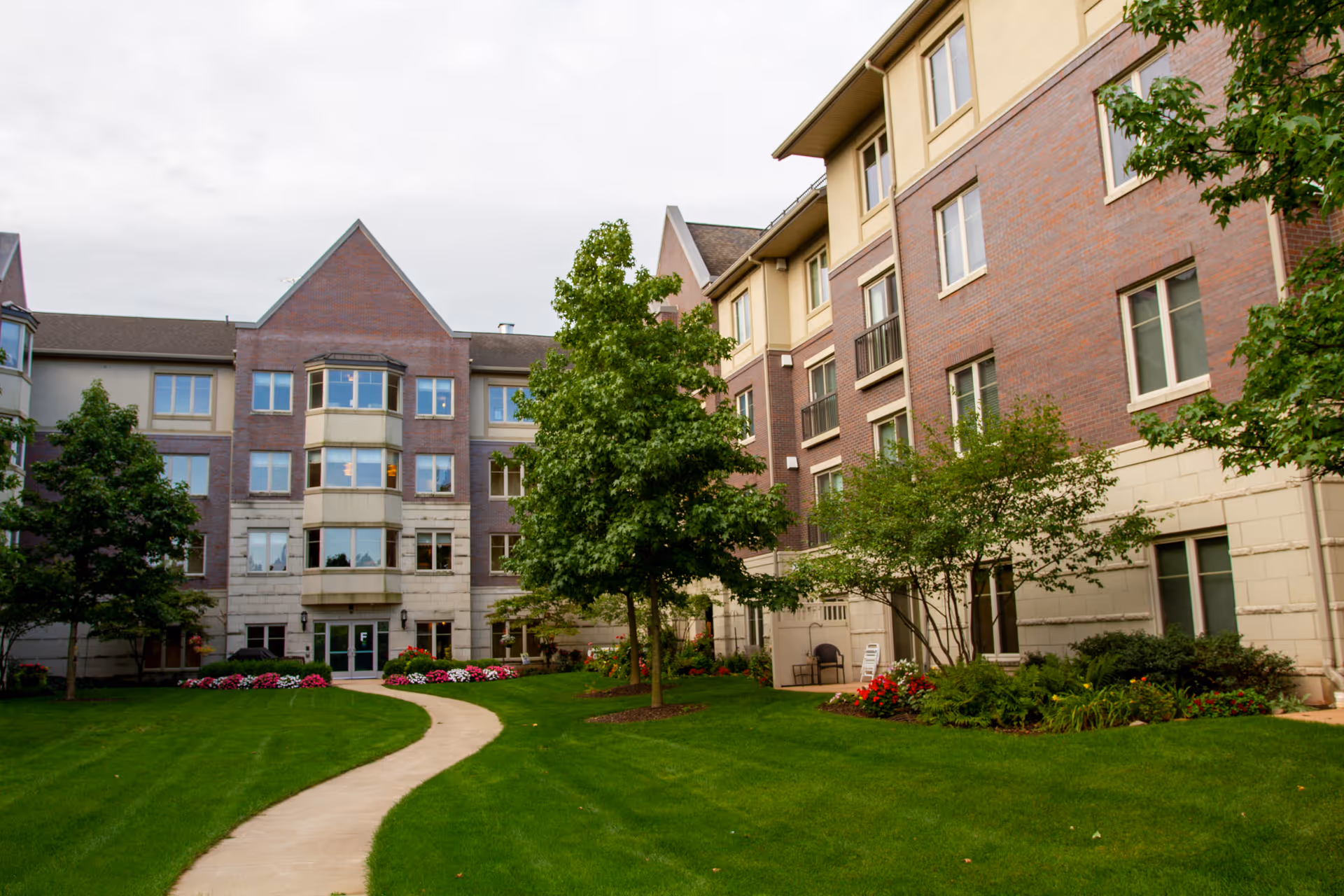 A well-maintained courtyard area of a senior living facility with a curved concrete pathway, green grass, trees, and flower beds. The multi-story building surrounding the courtyard has a brick and stone exterior with multiple windows.
