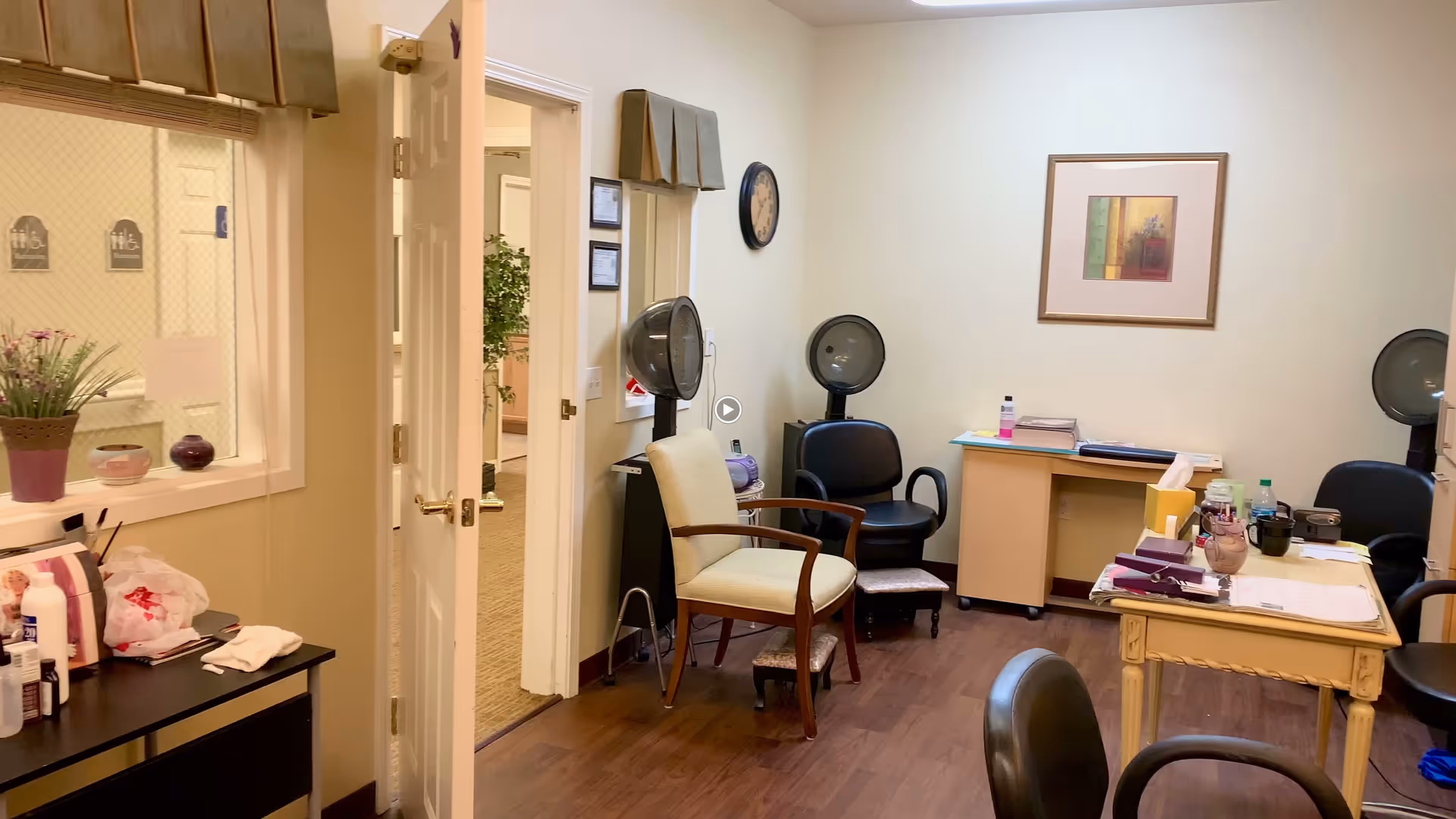 Interior view of a room in an assisted living facility with chairs, hair drying stations, a small desk with papers and bottles, a wall clock, and framed artwork. The room has wood flooring and light-colored walls. A door is open to a hallway with carpeted flooring and plants visible.