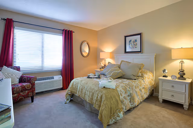A well-lit bedroom with a made bed, two nightstands with lamps, a patterned armchair, red curtains, and a window air conditioner.