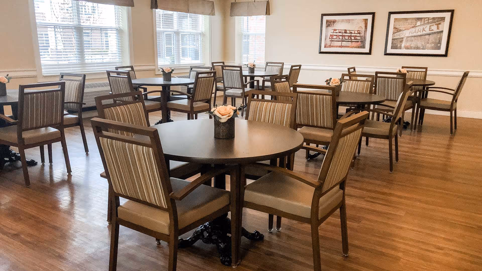Bright communal dining room with round tables, striped cushioned chairs, wood floors, windows and small floral centerpieces.