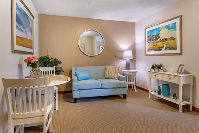 Cozy sitting area with a light blue loveseat, round wall mirror, side tables, framed artwork, and a small dining table in a neutral-toned room.