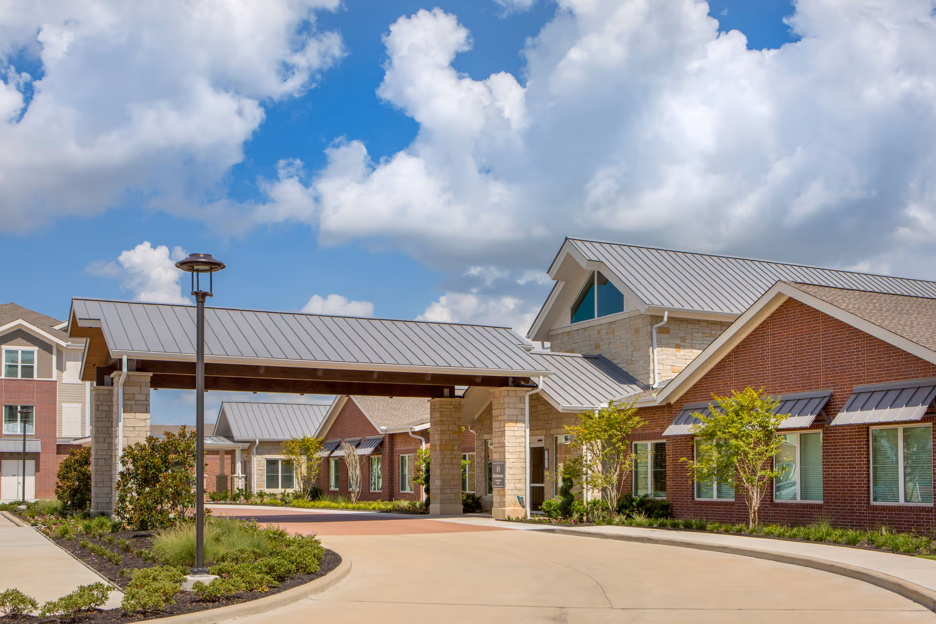 Exterior view of Delaney Parkway Lakes facility showing a covered entrance with stone pillars, brick and stone buildings with metal roofs, landscaped greenery, and a partly cloudy blue sky.