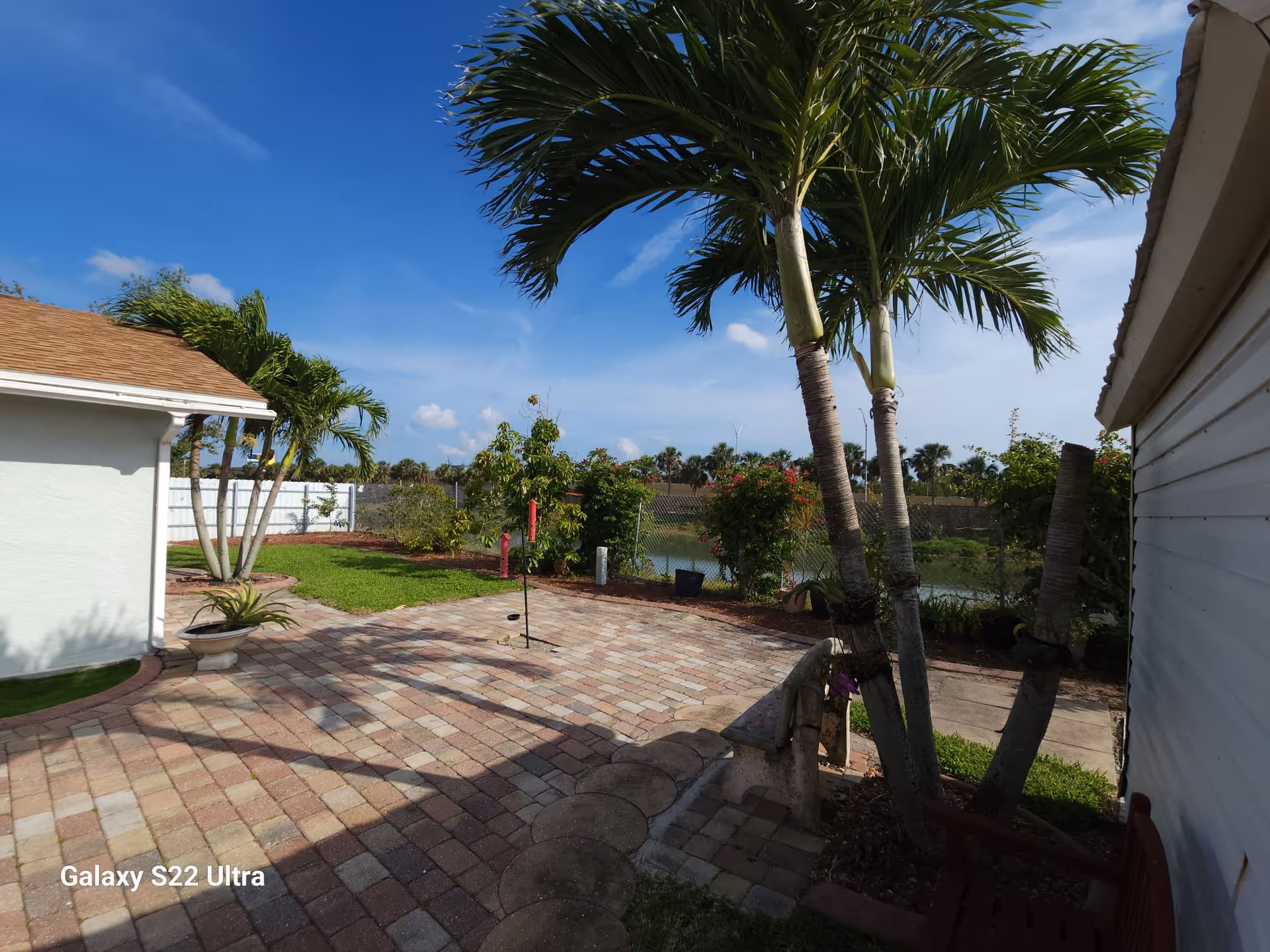 Outdoor patio area with brick pavers, palm trees, and green grass. There are some bushes and plants along the fence line, and a small body of water is visible in the background under a blue sky with a few clouds.