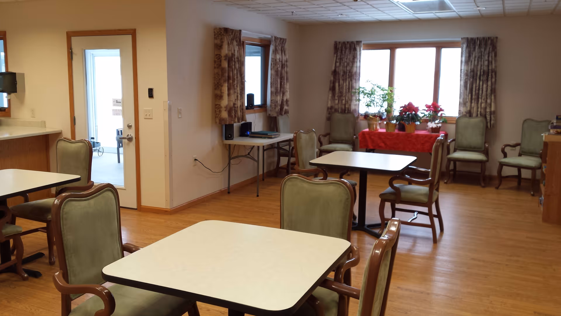 A dining area with several square tables and green cushioned chairs with wooden frames. There are large windows with patterned curtains, a table with potted plants and poinsettias covered with a red cloth, and a small table with electronic equipment against the wall. The floor is wooden, and the walls are light-colored.