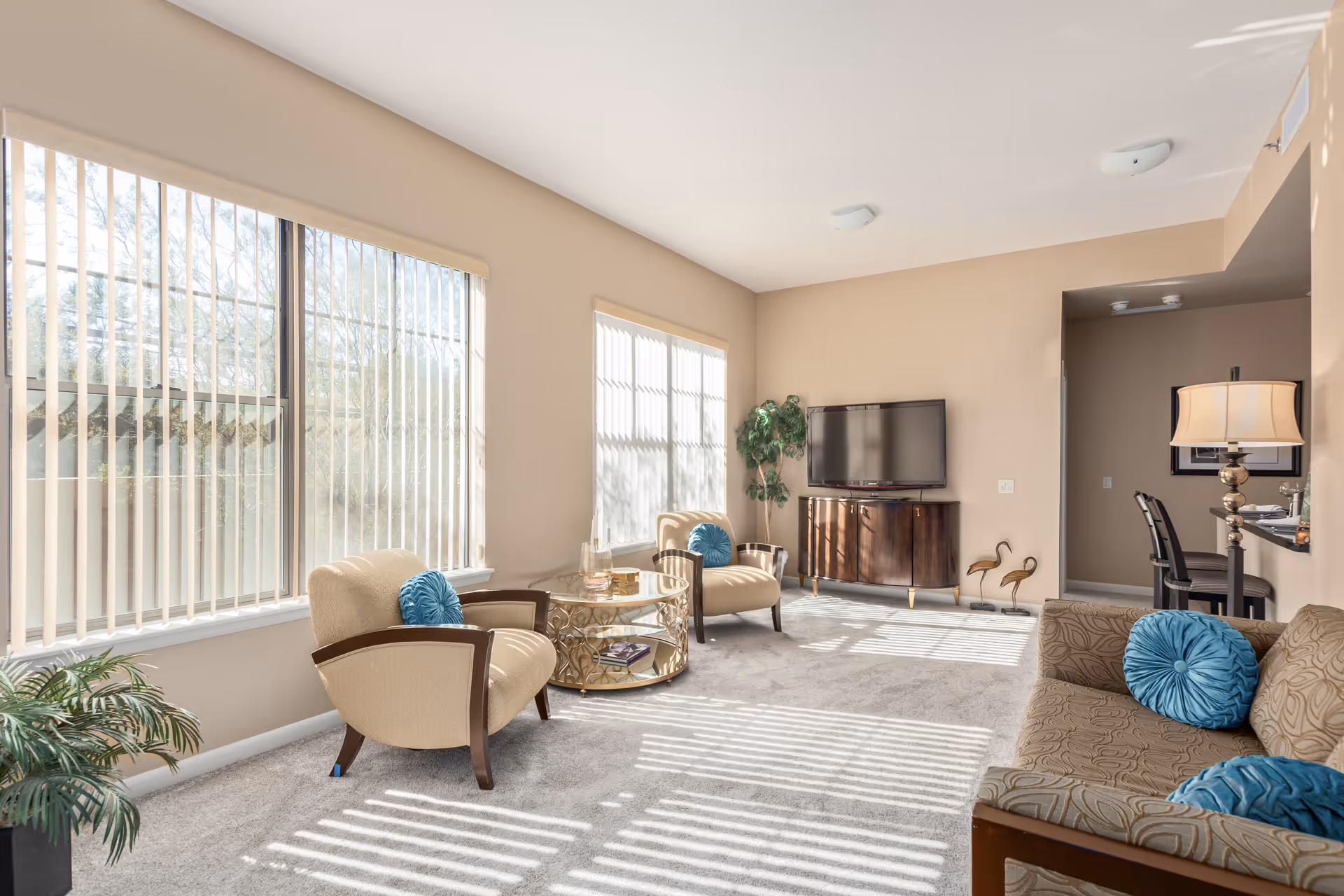 Bright furnished living room with armchairs, a sofa, a TV on a stand, a round coffee table, and large windows with vertical blinds.