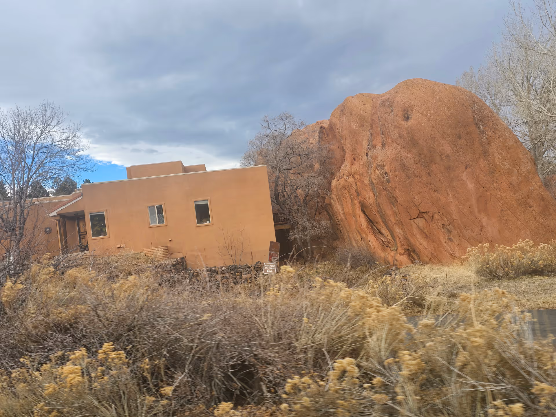 A tan-colored building with a flat roof next to a large reddish rock formation. The building has three windows visible and is surrounded by dry bushes and leafless trees under a cloudy sky.