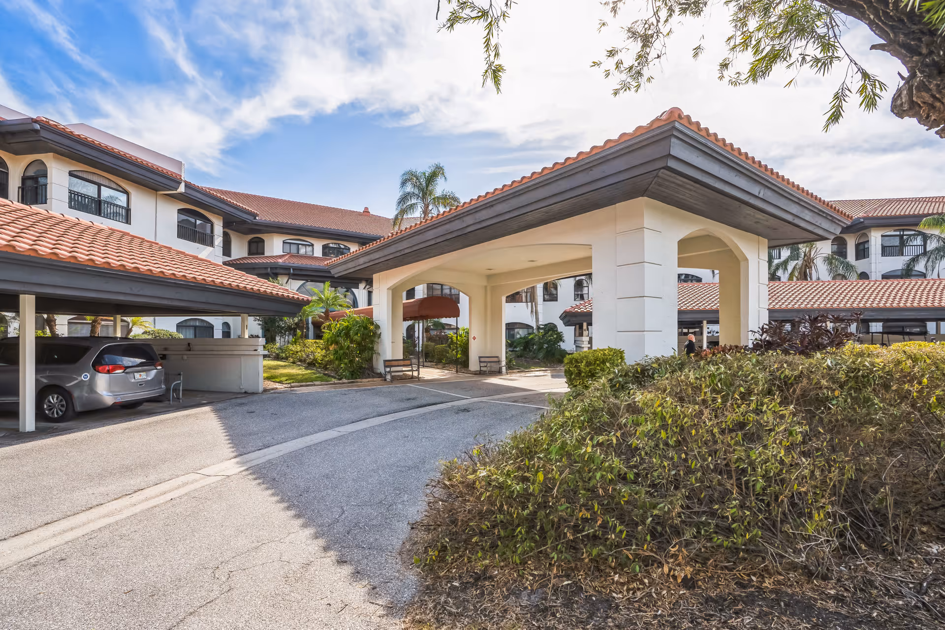 Exterior view of The Barclay at Sarasota senior living facility showing a covered entrance with arches, a driveway, parked cars under a carport, and a multi-story building with red-tiled roofs and balconies surrounded by greenery.