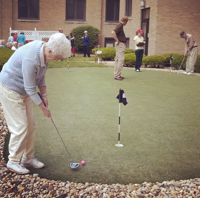 Elderly people playing mini golf on a putting green outside a brick building, with some people watching in the background.