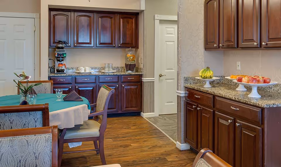 Interior view of a dining area in a senior living facility featuring a round table with a teal tablecloth and four chairs. The room has wooden cabinets with granite countertops, a coffee maker, cups, and plates on the counter. There are also two white pedestal fruit bowls with bananas and apples on the right countertop. The floor is wooden, and there are two white doors visible in the background.