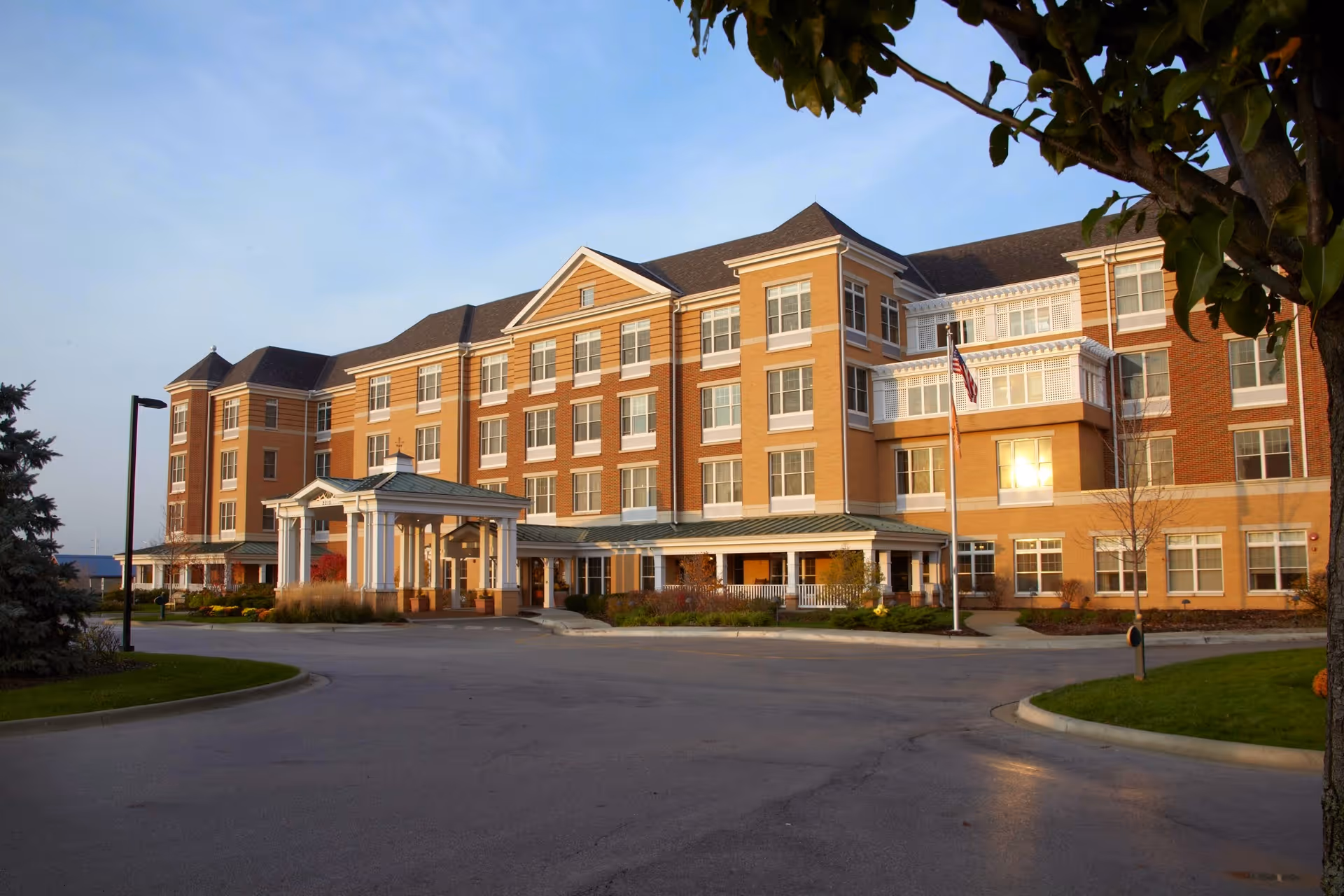 Exterior view of a large, multi-story senior living facility building with a covered entrance, multiple windows, and an American flag on a flagpole. The building is surrounded by a paved driveway and landscaped greenery under a clear sky.