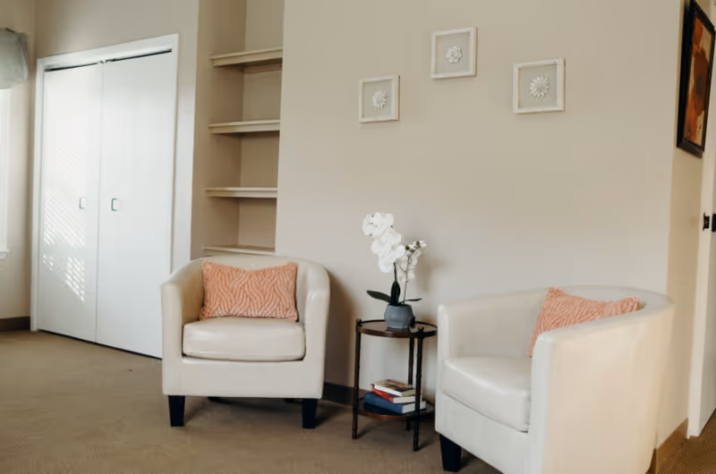 A cozy seating area with two white armchairs each with a coral-colored pillow, a small round wooden side table with a potted white orchid and some books, beige walls with three small framed floral artworks, and a white closet with sliding doors in the background.