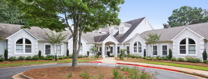 Exterior view of a single-story senior living facility building with white walls, multiple arched windows, a central entrance with a covered porch, surrounded by landscaped greenery and trees under a clear blue sky.