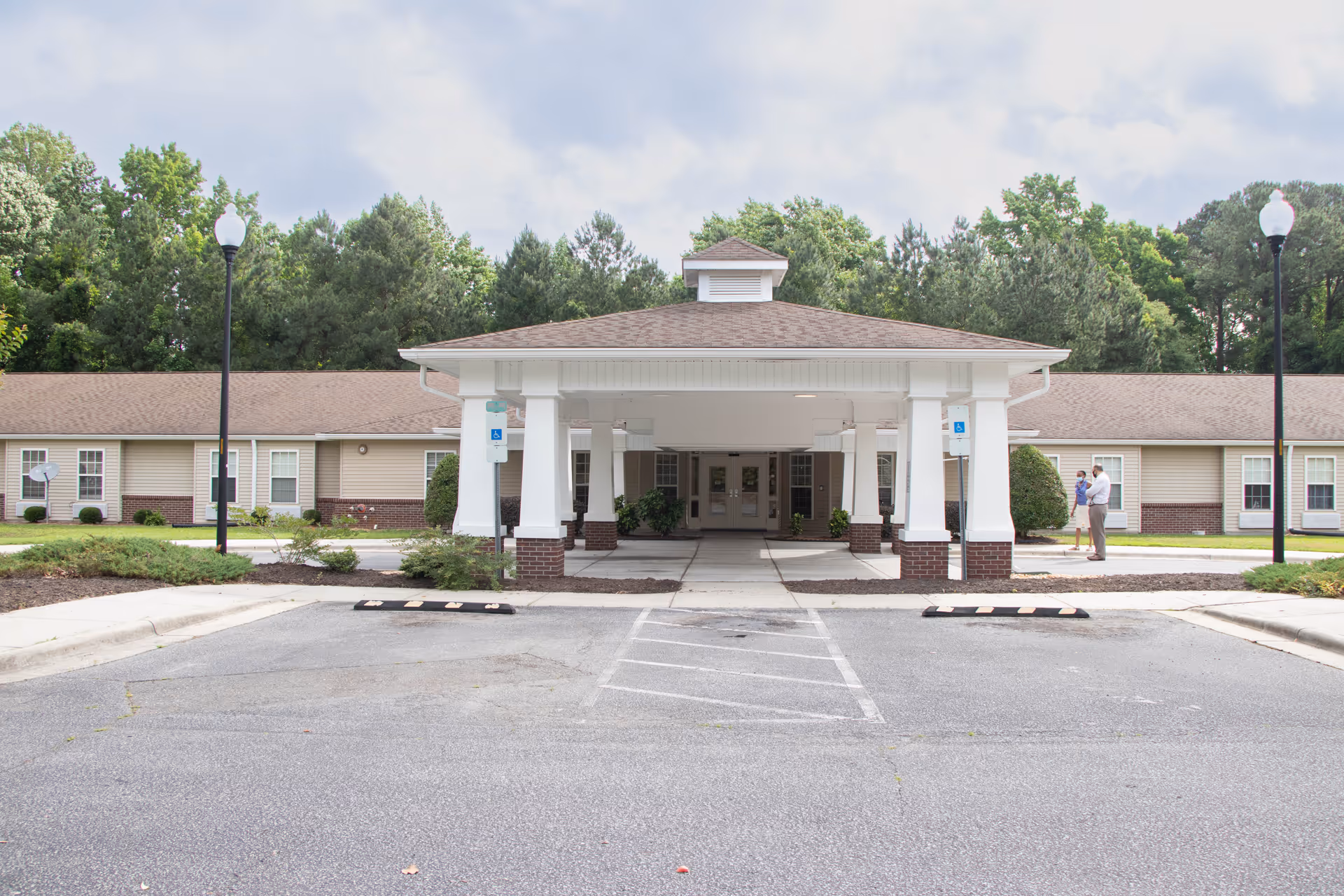 Front exterior view of Somerset Court of Goldsboro, showing a single-story building with a covered entrance supported by white columns with brick bases. There are two people standing near the entrance on the right side, and the area is surrounded by trees and greenery. The parking lot and handicap parking spaces are visible in the foreground.