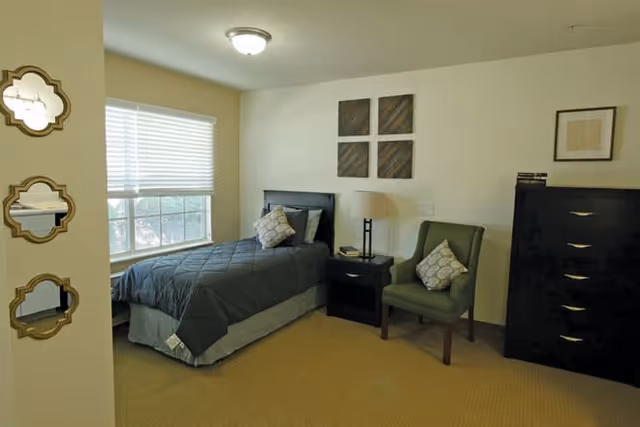 A bedroom with a single bed covered in a dark gray quilt and two pillows, a green upholstered chair with a patterned pillow, a black nightstand with a lamp, a black chest of drawers, and three decorative wall mirrors on the left wall near a window with blinds.