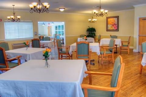 Sunlit dining room with round tables, white tablecloths, and wooden chairs in a senior facility.