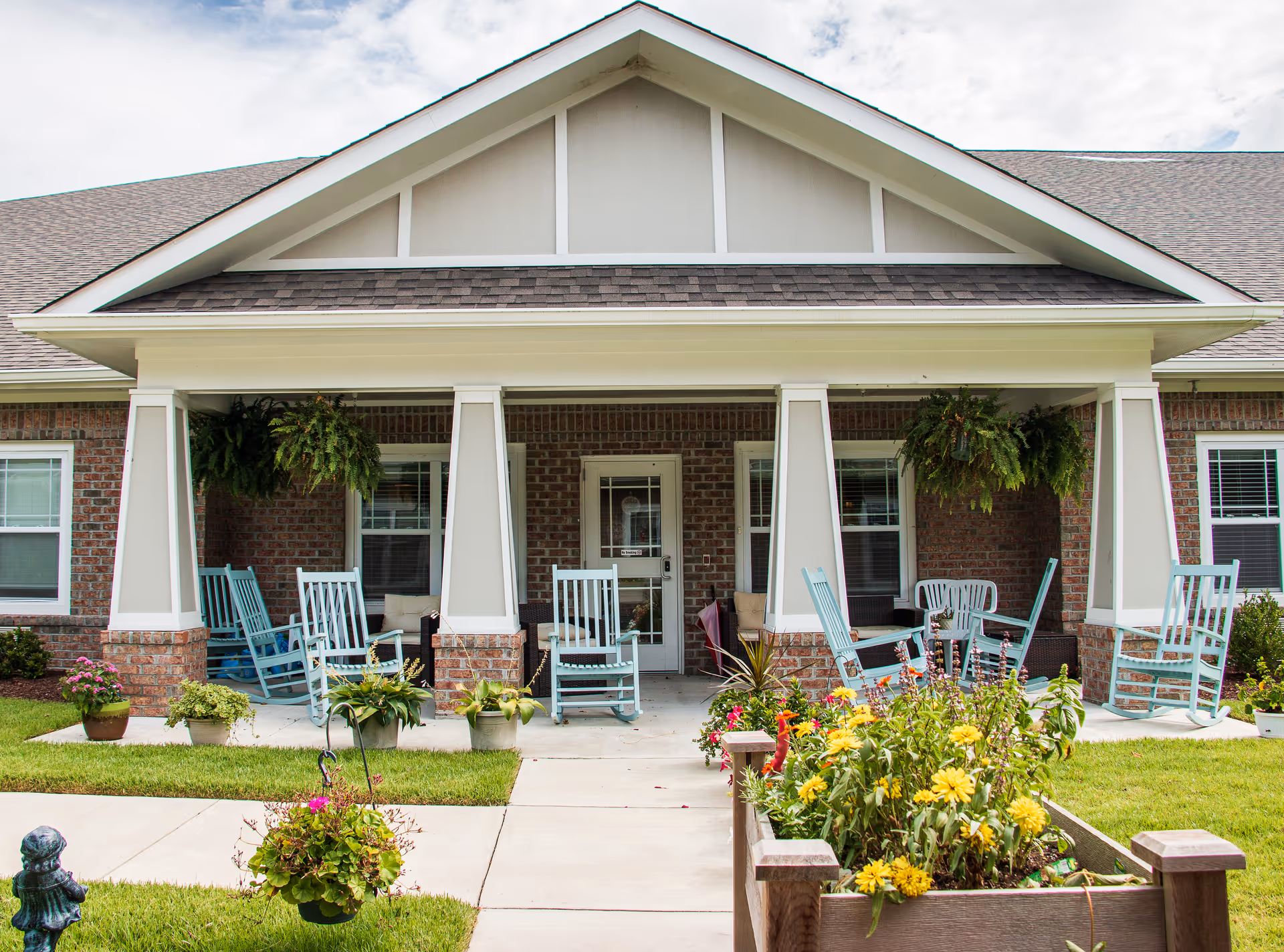 Covered front porch of a residential building with light-blue rocking chairs, hanging ferns, and flower planters.