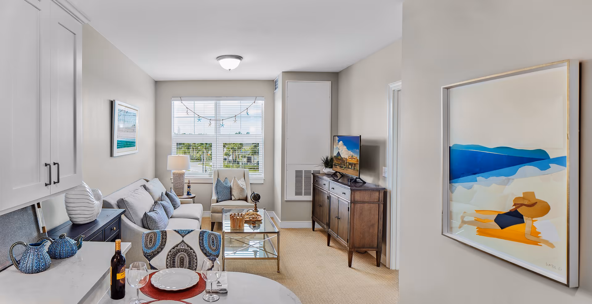 A bright and cozy living room area with a gray sofa, an armchair, a glass coffee table, and a wooden TV stand with a flat-screen TV. The room has a large window with blinds and a string of starfish decorations. There is a kitchen counter with a bottle of wine, two wine glasses, and a plate with cutlery in the foreground. The walls are light-colored, and there is a framed beach-themed artwork on the right wall.