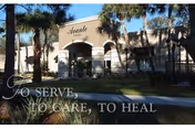 Exterior view of Avante at Ocala building with palm trees and landscaping in front. The building has an arched entrance and beige walls. Text overlay at the bottom left reads 'TO SERVE, TO CARE, TO HEAL'.
