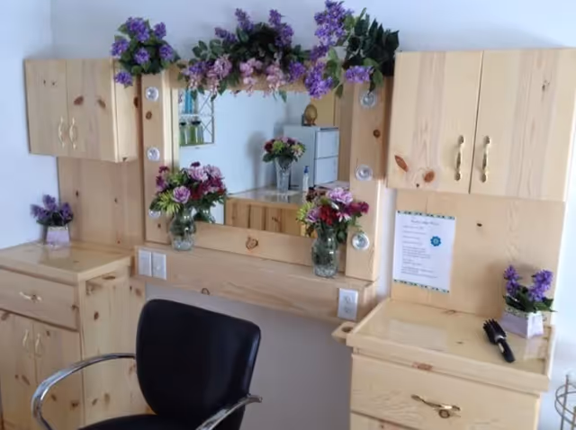 A salon-style wooden vanity with a mirror decorated with purple flowers, vases of blooms, cabinets, and a black styling chair.