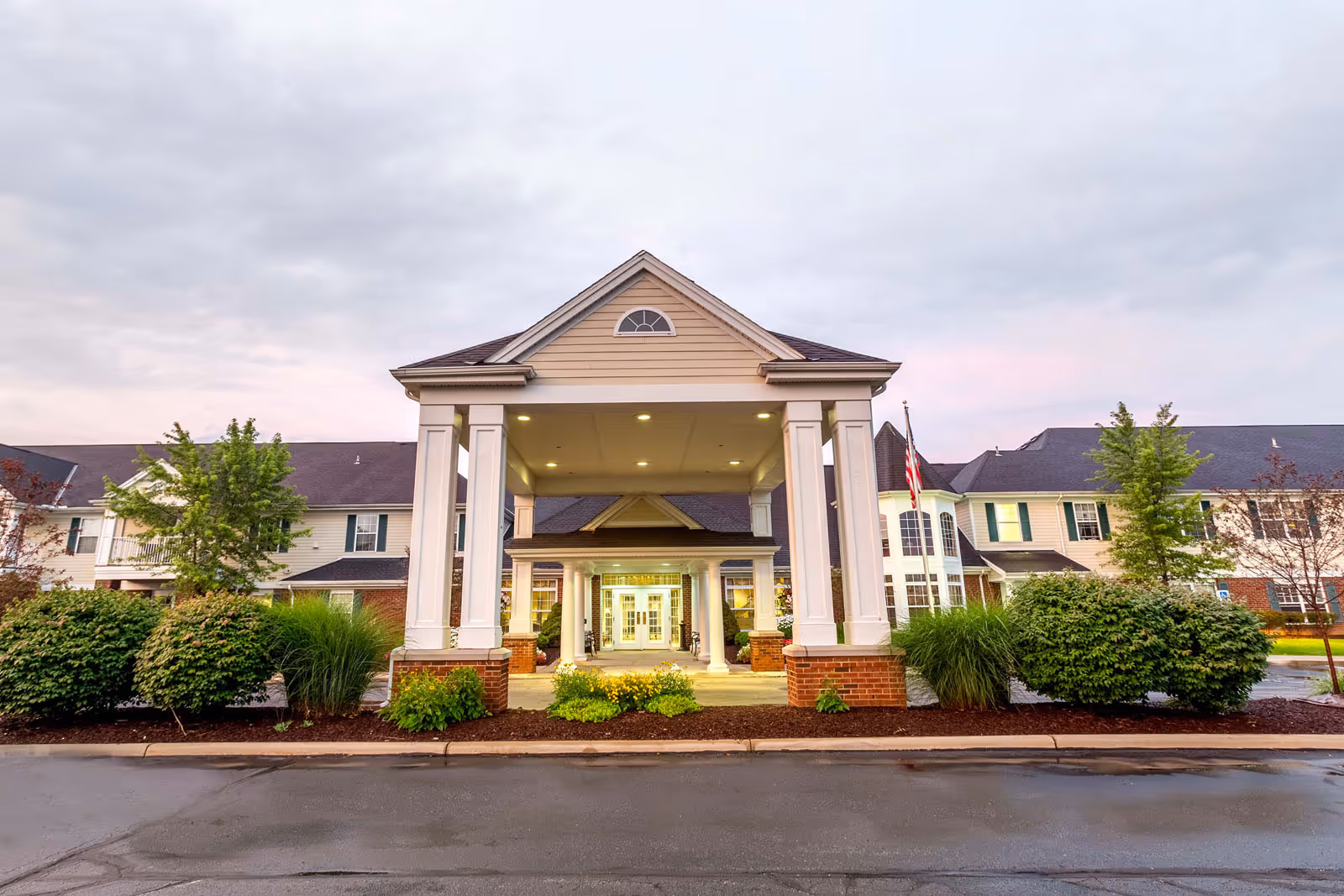 Front exterior view of Independence Village of Aurora, showing a large covered entrance with white columns and brick bases, surrounded by landscaped bushes and trees under a cloudy sky.