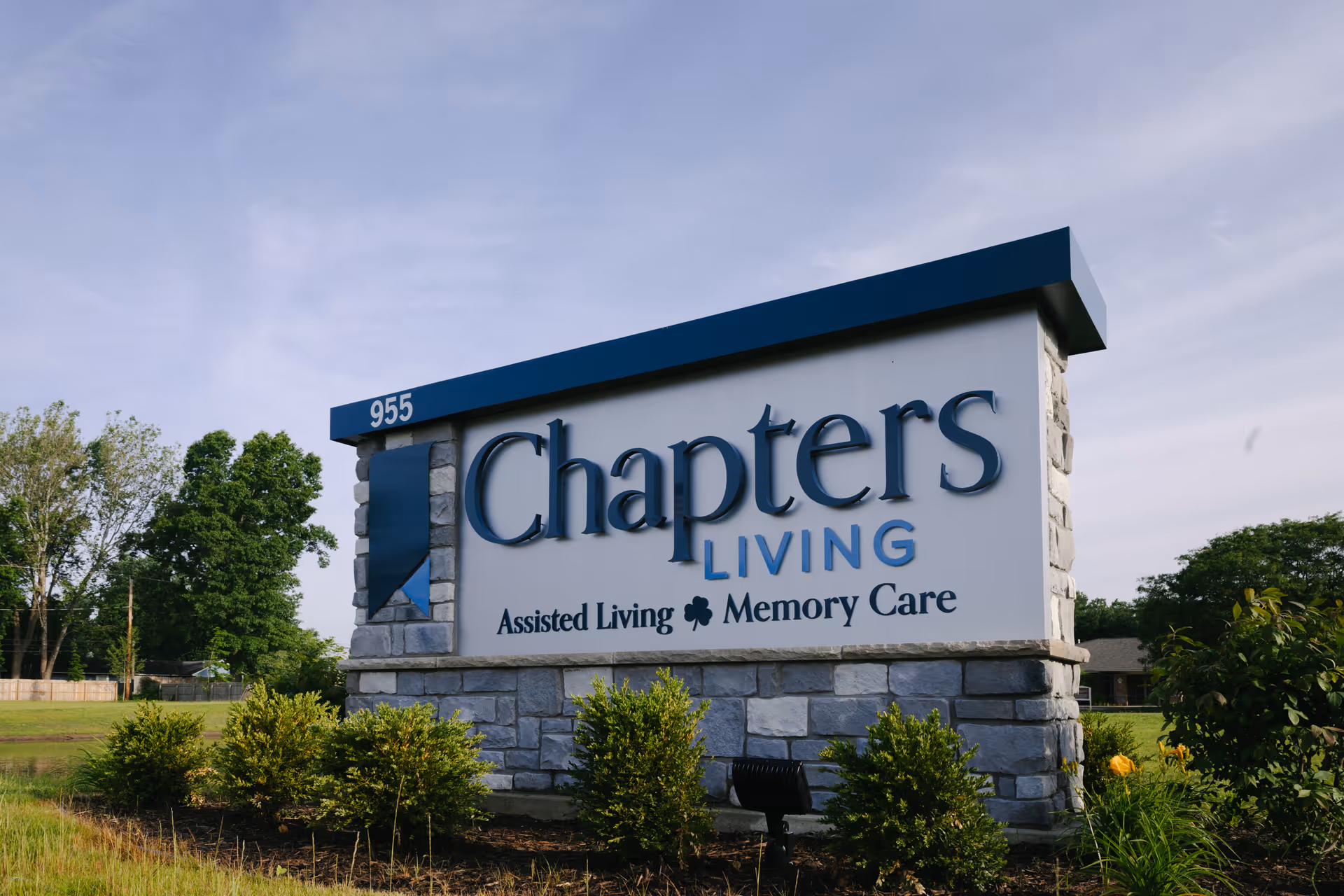 Outdoor stone sign for Chapters Living, an assisted living and memory care facility, with greenery and trees in the background under a partly cloudy sky.