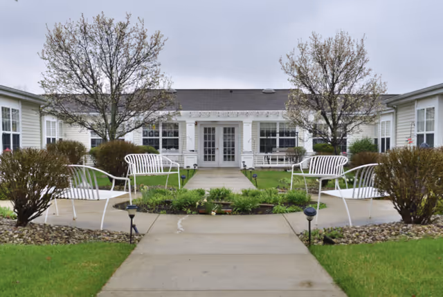 A paved courtyard with white metal benches and a central walkway leading to the entrance of a single-story building, flanked by small trees and shrubs.