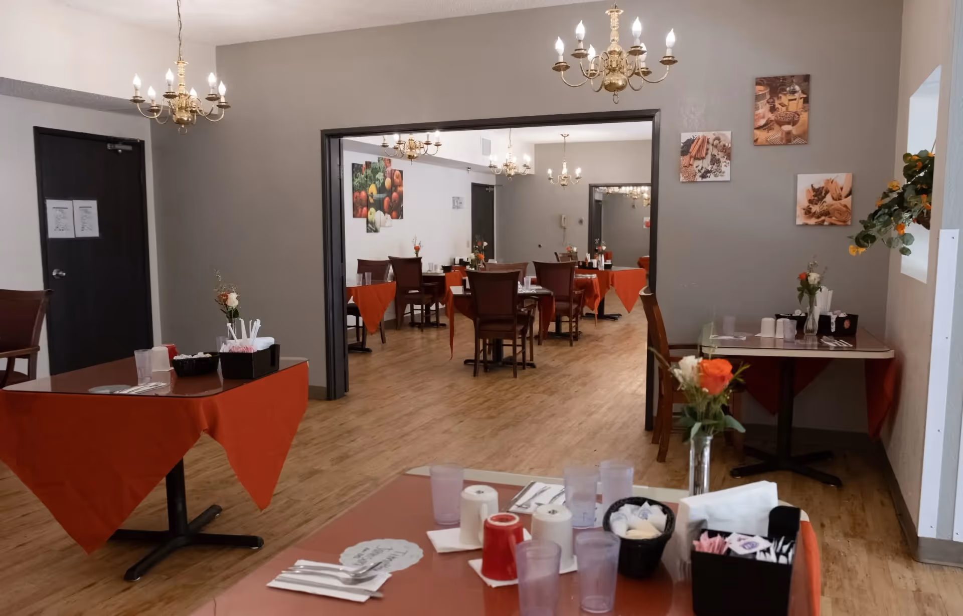 Interior view of a dining room in Downey Retirement Center with multiple tables covered with red tablecloths. Each table is set with utensils, cups, napkins, and small flower vases. The room has wooden flooring, beige walls with framed pictures, and brass chandeliers hanging from the ceiling.