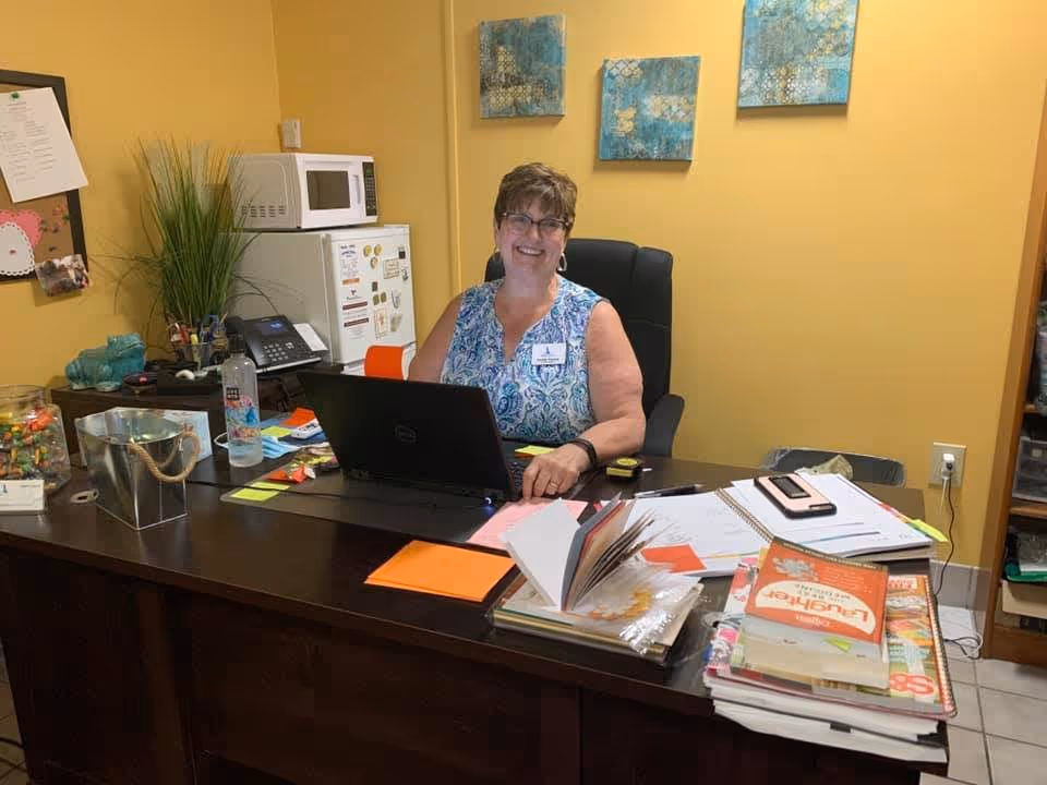 A woman sitting at a desk in an office environment, smiling at the camera. The desk is cluttered with papers, notebooks, a laptop, and office supplies. Behind her is a small refrigerator with a microwave on top, a bulletin board, and some wall art on a yellow wall.