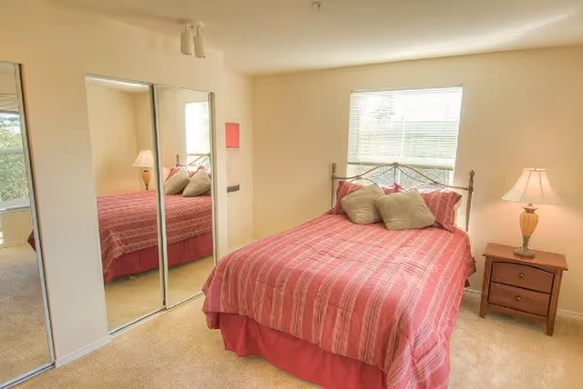A bedroom with a bed covered in a red and pink striped bedspread and several pillows. There is a wooden nightstand with a lamp on the right side of the bed. A window with blinds is behind the bed, and a mirrored closet door is on the left side of the room. The walls are light-colored and the floor is carpeted.