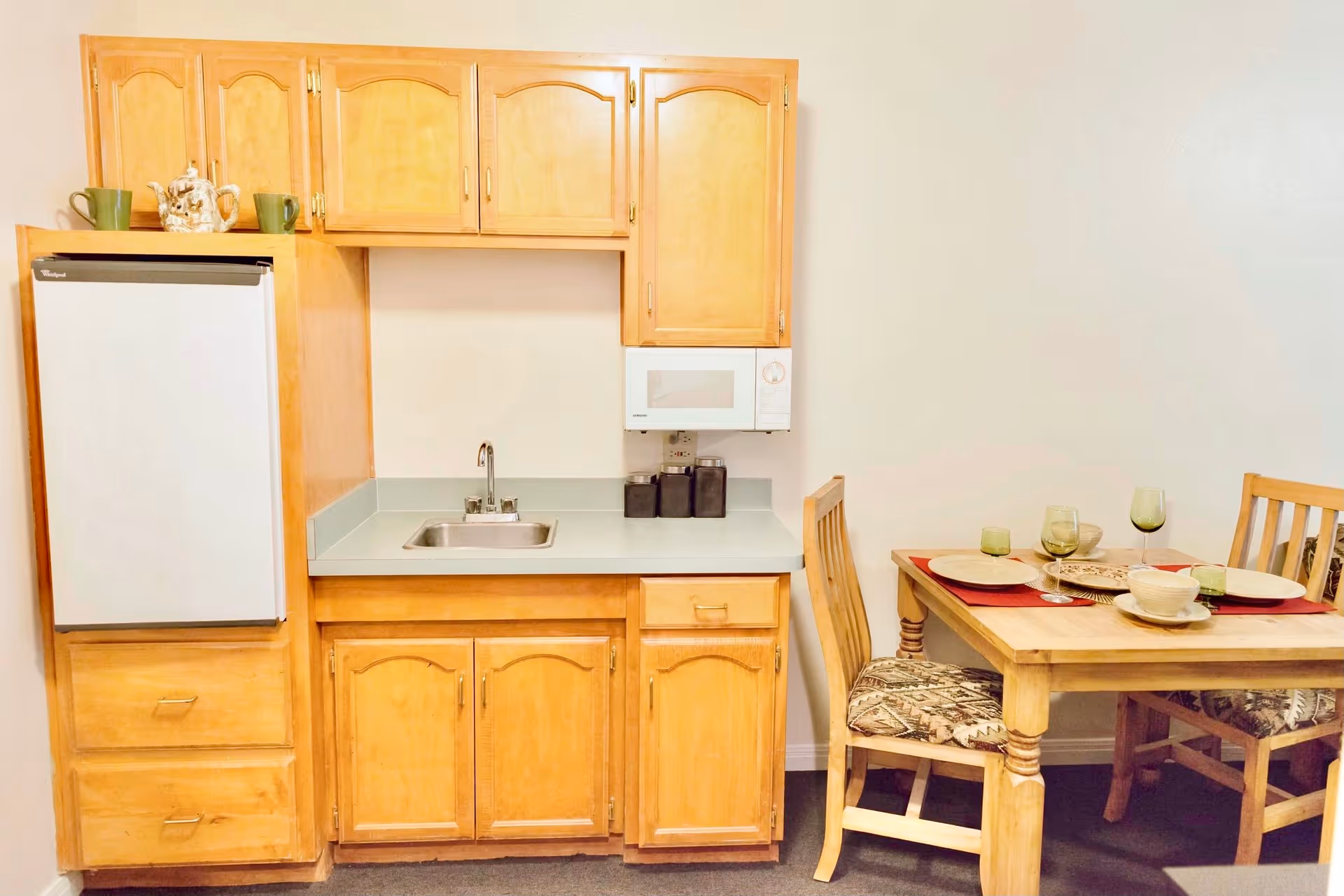 Small kitchen area with light wood cabinets, a white refrigerator, a sink, a microwave, and a wooden dining table set with four chairs and place settings including plates, bowls, and glasses.