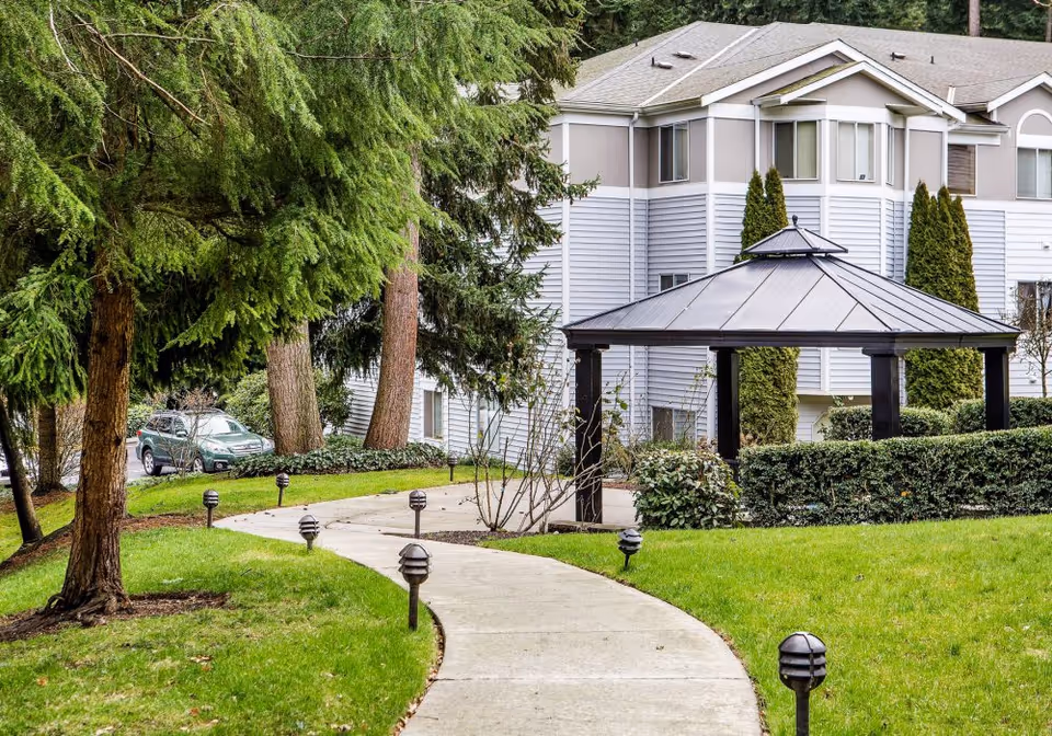 A winding concrete pathway lined with small black outdoor lights leads through a green lawn with trees and shrubs toward a gazebo with a metal roof. In the background, there is a multi-story residential building with gray and white siding and several windows.