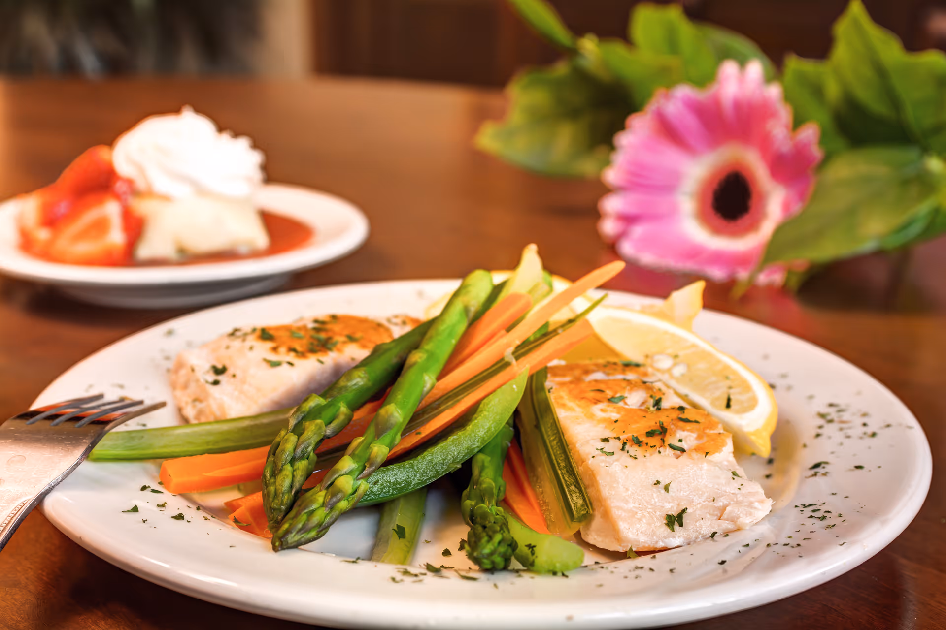 A plated meal of seared fish with asparagus, carrots and lemon on a white plate with a fork, a dessert plate and a pink flower blurred in the background.
