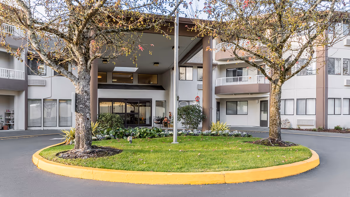 Entrance area of Capital Place Retirement Community showing a circular driveway with a grassy island in the center, two trees, and a flagpole. The building has multiple floors with balconies and large windows, and the entrance has a covered drop-off area.