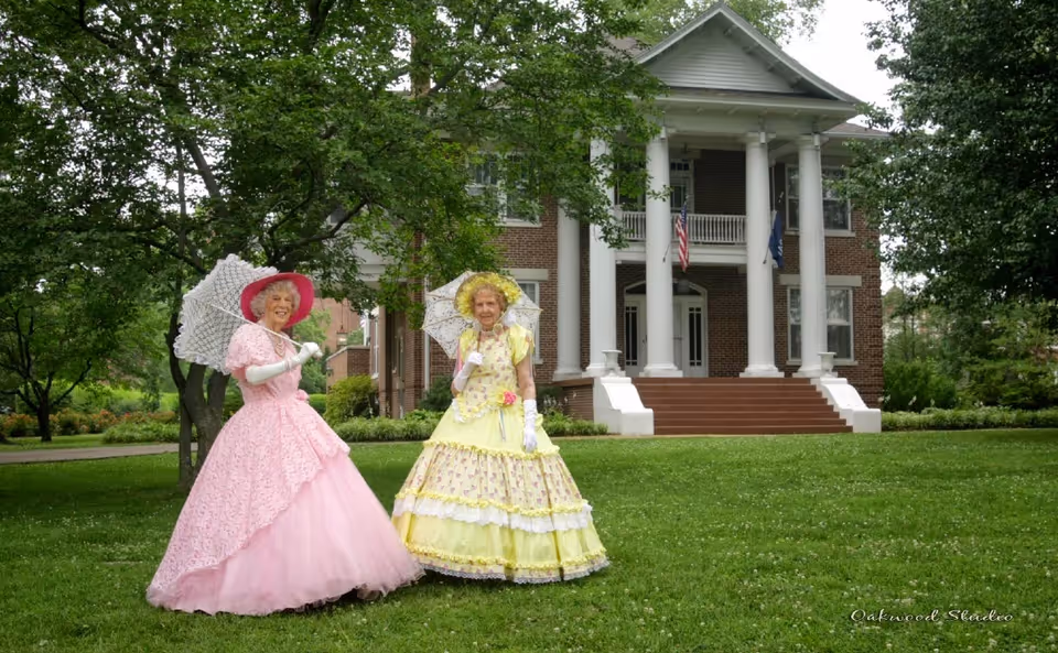 Two women in Victorian-style dresses holding parasols stand on the lawn in front of a large columned brick building.