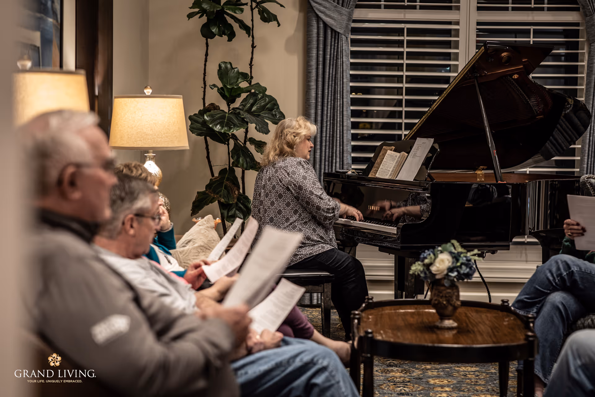 A group of elderly people sitting in a cozy living room, holding sheets of paper, while a woman plays a grand piano near a window with closed blinds. The room is warmly lit with a lamp and decorated with a tall plant and a small table with a flower arrangement.