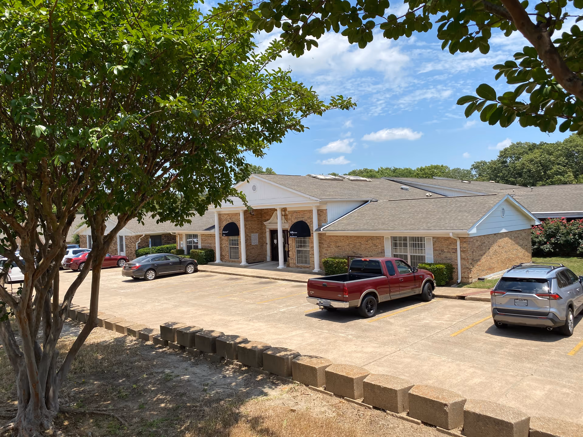 Front exterior of a single-story brick senior living building with white columns, parked cars, and trees in the foreground under a blue sky.