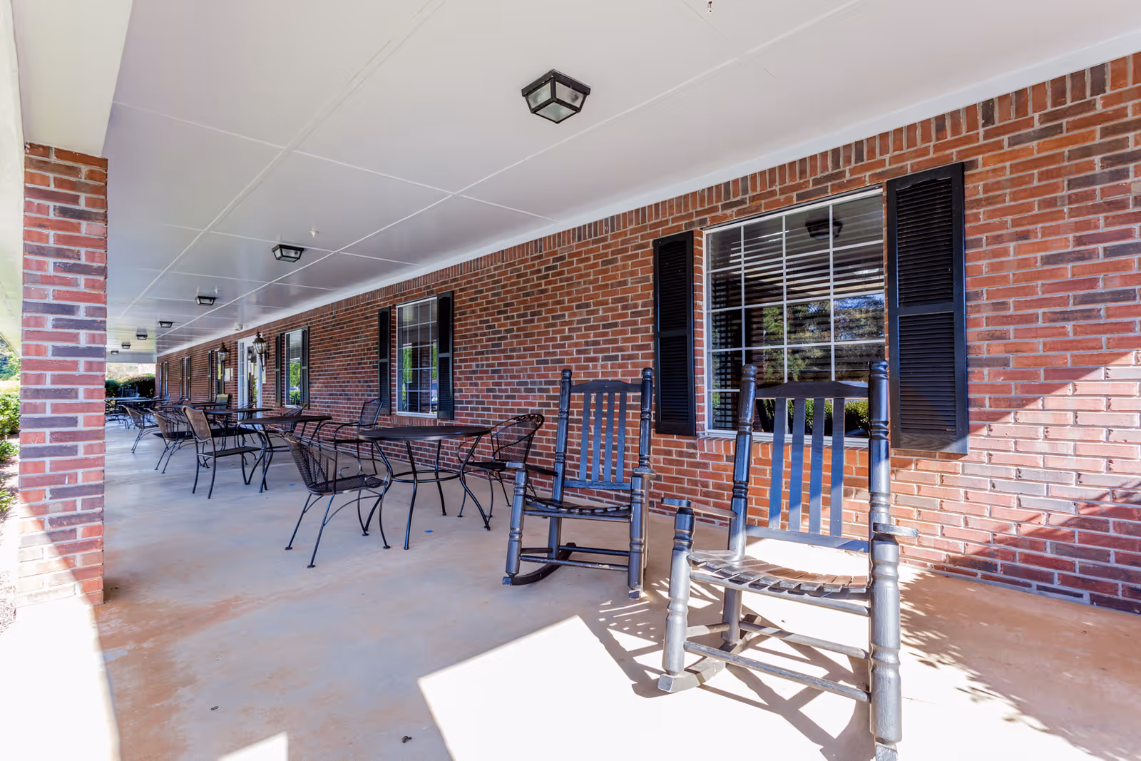 Covered outdoor patio area at Park Place Nursing and Rehab Facility with brick walls, black rocking chairs, metal tables and chairs, and windows with black shutters.