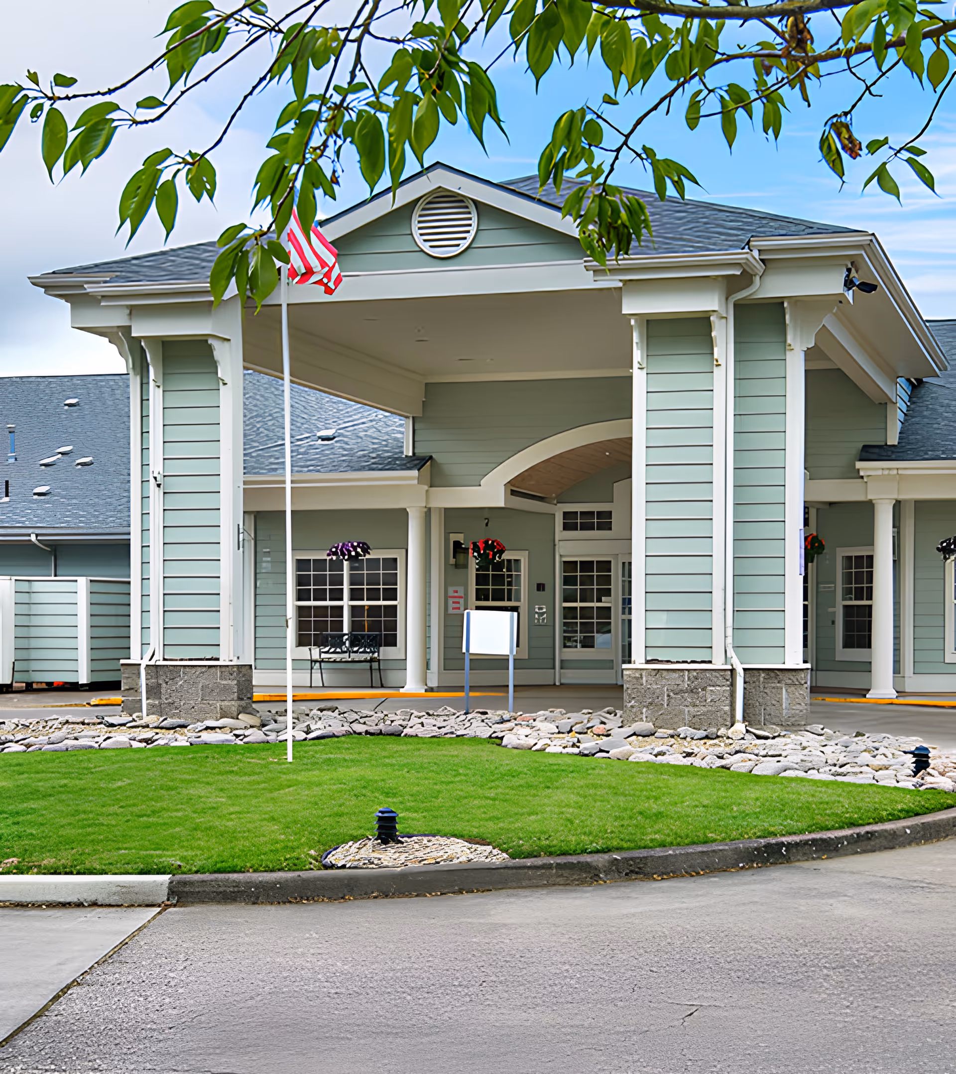Front entrance of the Spring Valley Assisted Living building with a covered porte-cochere, American flag, and landscaped lawn.