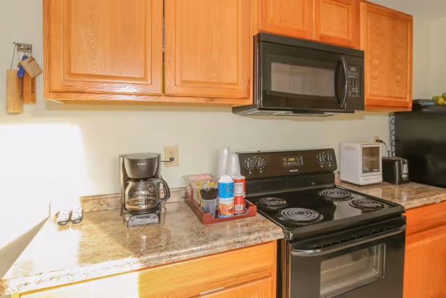 A kitchen countertop with wooden cabinets, a coffee maker, condiments on a tray, a black electric stove with coil burners and an over-the-range microwave.