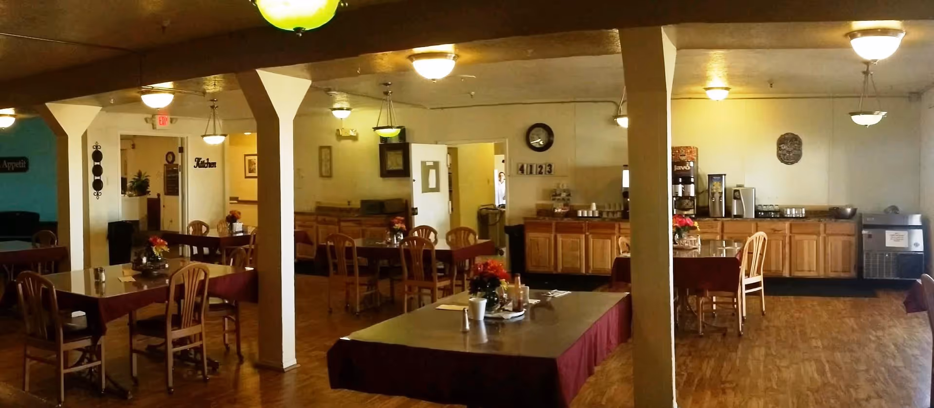 Interior view of a dining room in Camino Retirement Apartments featuring several tables with chairs, each table decorated with a small flower arrangement. The room has wooden flooring, multiple ceiling lights, and a counter area with coffee machines and cups. There are white pillars dividing the space and a clock on the wall showing the time as 4:12:3.