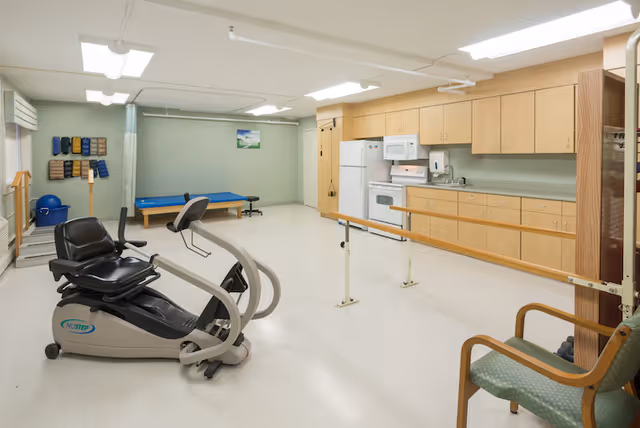 A rehabilitation room with exercise equipment including a recumbent bike, parallel bars for walking practice, a treatment table, and a kitchenette with wooden cabinets, a refrigerator, microwave, and sink. The room has light green walls and bright overhead lighting.
