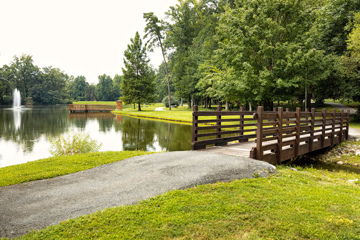 A wooden footbridge crossing a small stream toward a pond with a fountain, surrounded by grassy banks and trees.